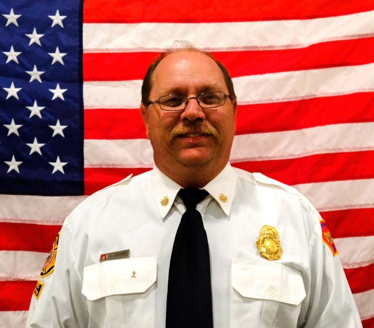 A middle-aged man in a white firefighter uniform with badges, smiling in front of an american flag.