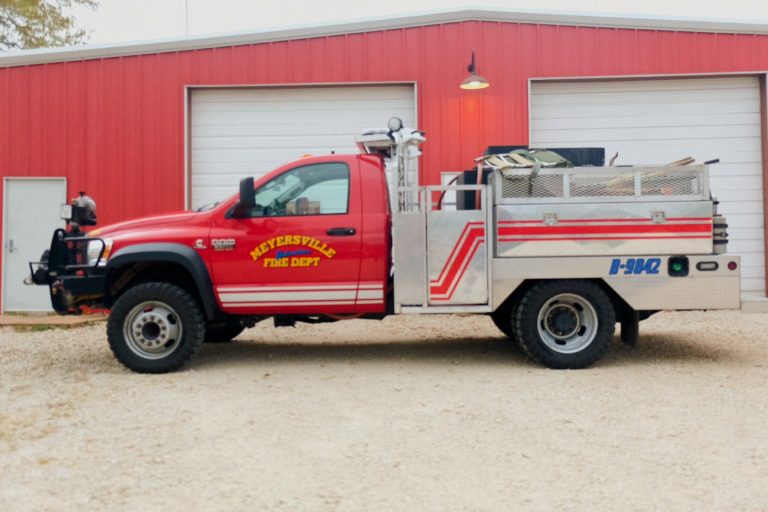 A red fire department utility truck parked outside a building with a red exterior wall. the truck is equipped with various firefighting tools and labeled "myersville fire dept.