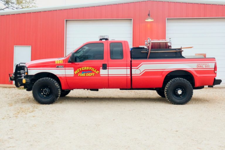 A red fire department pickup truck with the label "watersville fire dept" parked in front of a gray building, equipped with emergency lights and firefighting tools on its bed.