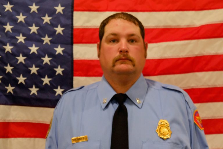 A portrait of Captain Andrew Venables in blue uniform, with a badge and name tag, standing in front of an american flag.