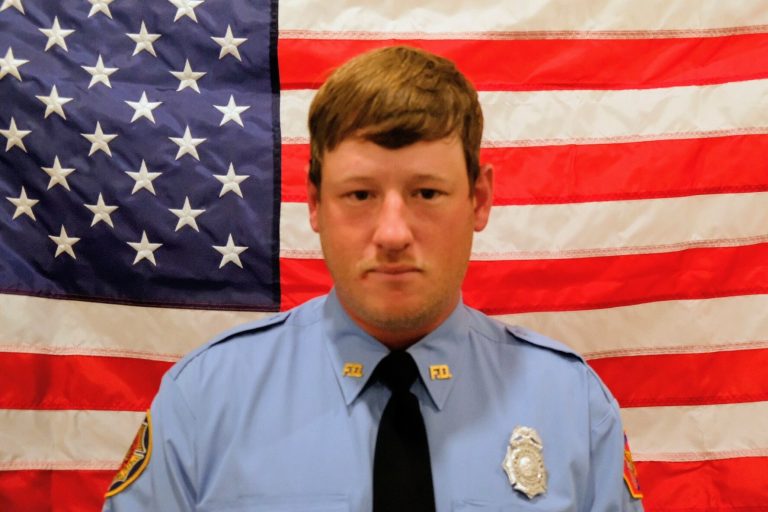 A police officer in a blue uniform, featuring a badge and patches, poses solemnly in front of an american flag.