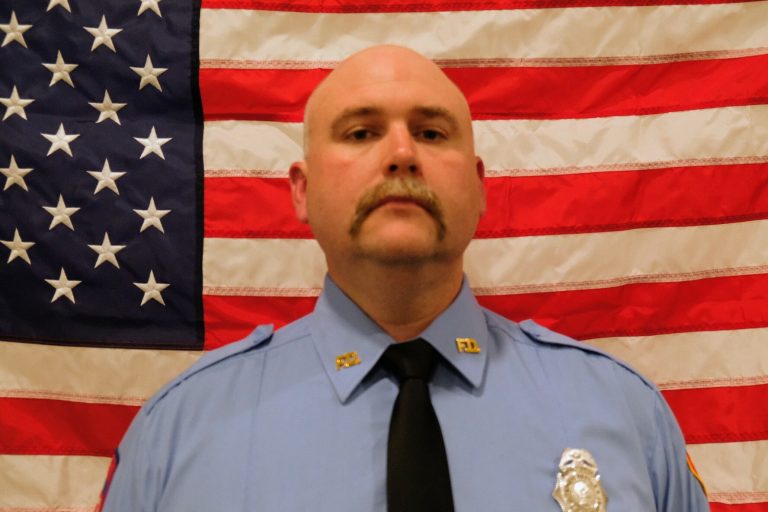 Darreck Kenjura Lieutentant with a serious expression, wearing a blue uniform with badges, stands in front of an american flag.