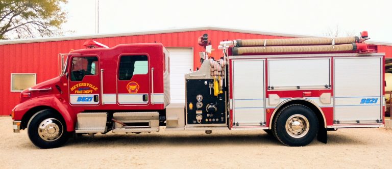 A red fire truck parked in front of a building with a firefighter standing at the rear, preparing equipment. the truck has distinguishing emblems and hoses visible on its side.