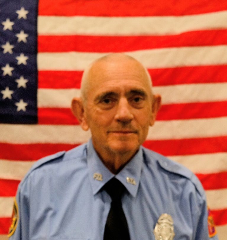 An elderly male police officer in uniform, with a serious expression, sitting in front of an american flag.