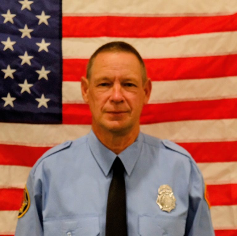 Firefighter Alan Winkelmannwith a badge and tie, stands in front of an american flag. he has a serious expression.