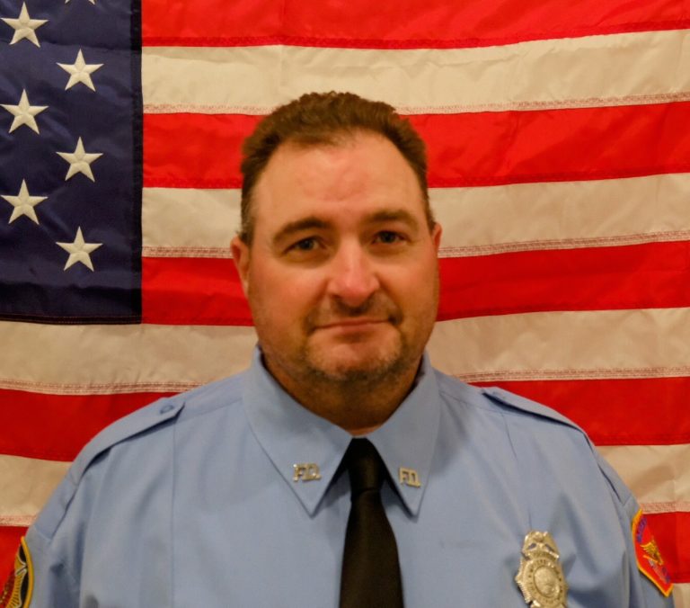 A portrait of a caucasian male firefighter in a blue uniform with badges, smiling slightly in front of an american flag.