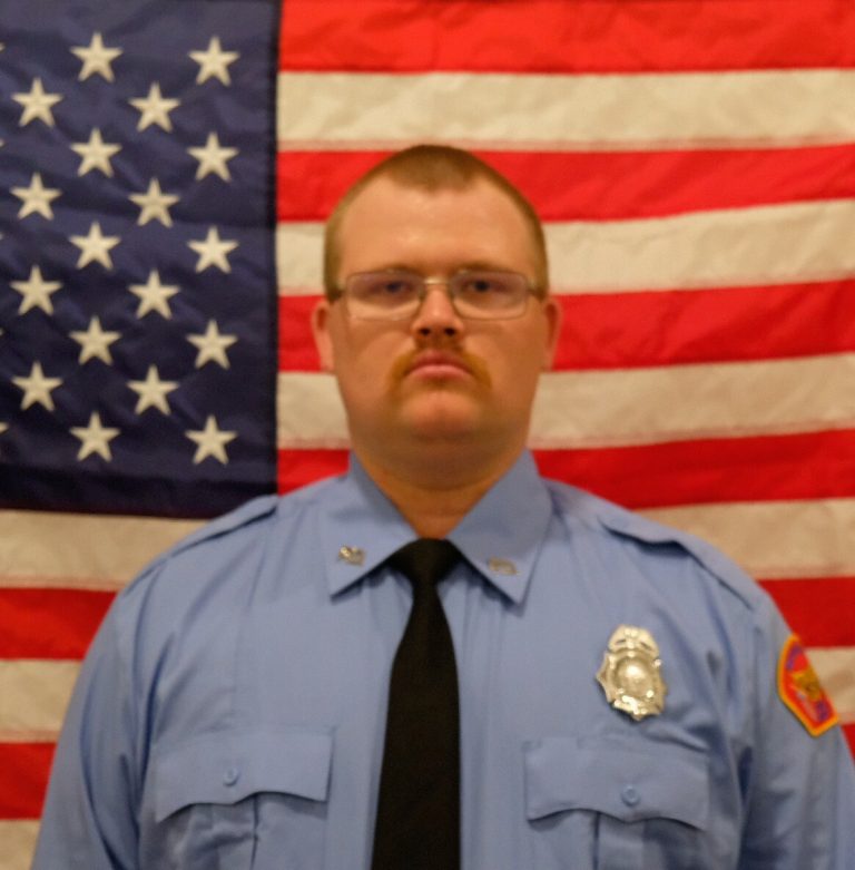 Jacob Tieman a uniformed firefighter stands solemnly in front of an american flag, wearing glasses and a serious expression. his badge is visible on his light blue shirt.