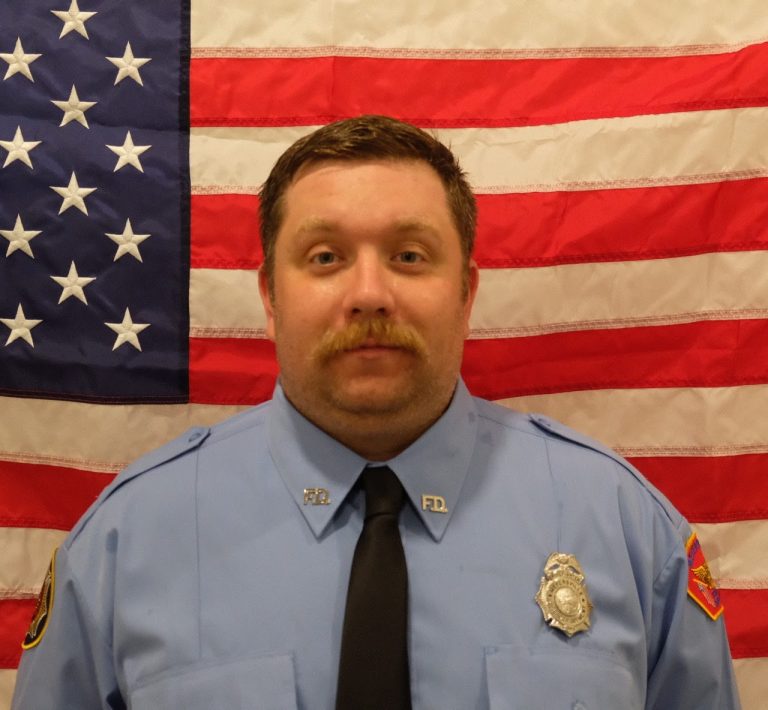 Firefighter Matt Martin in a blue firefighter uniform with badges, standing in front of an american flag. he has a mustache and appears serious.
