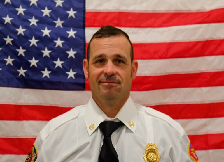 A man in a white uniform with a badge, smiling slightly, sits in front of an american flag, displaying a professional demeanor.