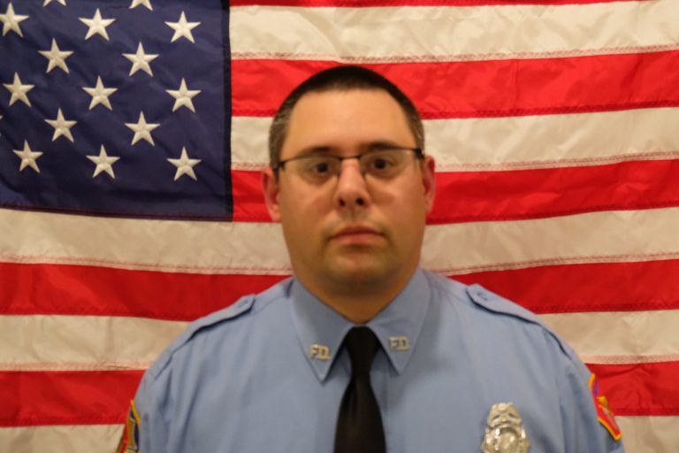 Matt Metzger Firefighter in a blue uniform with a badge, standing in front of an american flag, looking directly at the camera. the focus is slightly blurred.