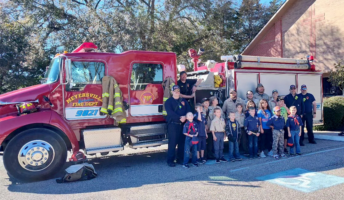 A group of children and adults posing happily with firefighters in front of a red fire truck outside a brick building on a sunny day.
