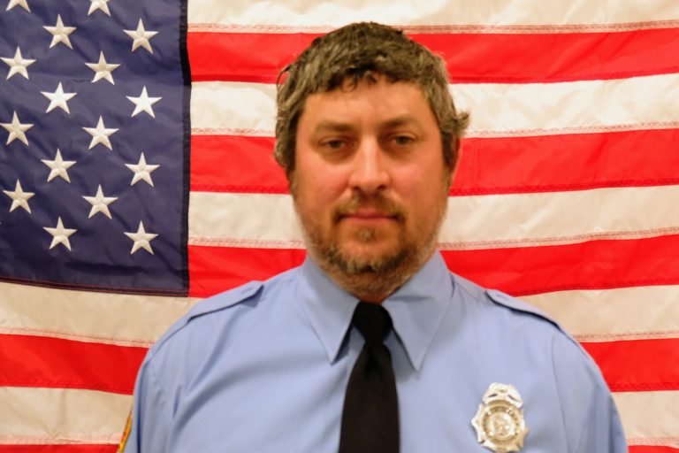 Rallin Wellmann, Firefighter in a blue uniform and badge stands in front of an american flag. he has a slight smile and is wearing a winter hat.