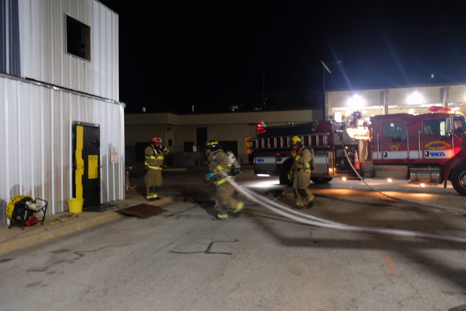 Firefighters in gear walk near a fire truck with hoses outside a building at night during an emergency response scene.