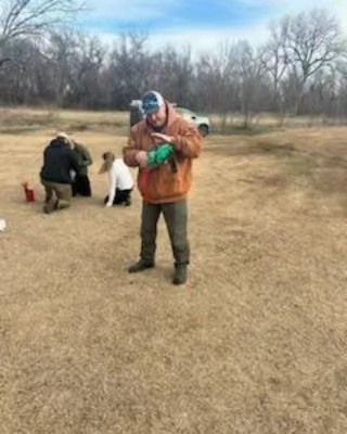 A man in a cap and orange sweatshirt opens a green bag in a grassy field. in the background, a group of people sit near red buckets, engaged in an activity.