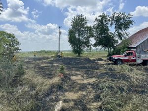 Three people stand near a telegraph pole in a field with patches of burnt grass. A red emergency vehicle is parked near a barn with a rusted roof. Trees and cloudy skies are visible in the background. Yellow tape sections off part of the scorched area.
