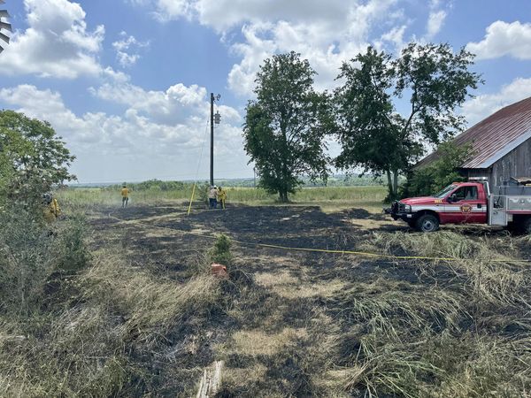 Three people stand near a telegraph pole in a field with patches of burnt grass. A red emergency vehicle is parked near a barn with a rusted roof. Trees and cloudy skies are visible in the background. Yellow tape sections off part of the scorched area.