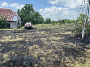 A small rural grass fire scene shows charred ground with blackened grass. A fire truck is parked near an old, weathered barn with a rusted roof on the left side. A firefighter in yellow gear is visible in the background, assessing the area under a partly cloudy sky.