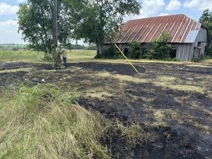 A person stands near a power pole in a field that shows signs of recent fire damage, with burnt ground and patches of blackened grass. A weathered, wooden barn with a rusty roof sits in the background, surrounded by green trees and vegetation. The sky is partly cloudy.