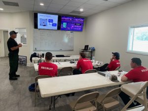 A man stands and presents to four seated individuals wearing red 'RECRUIT' shirts in a classroom setting. The room has a whiteboard with notes, and multiple screens displaying various images and information. The recruits listen attentively, facing the instructor.