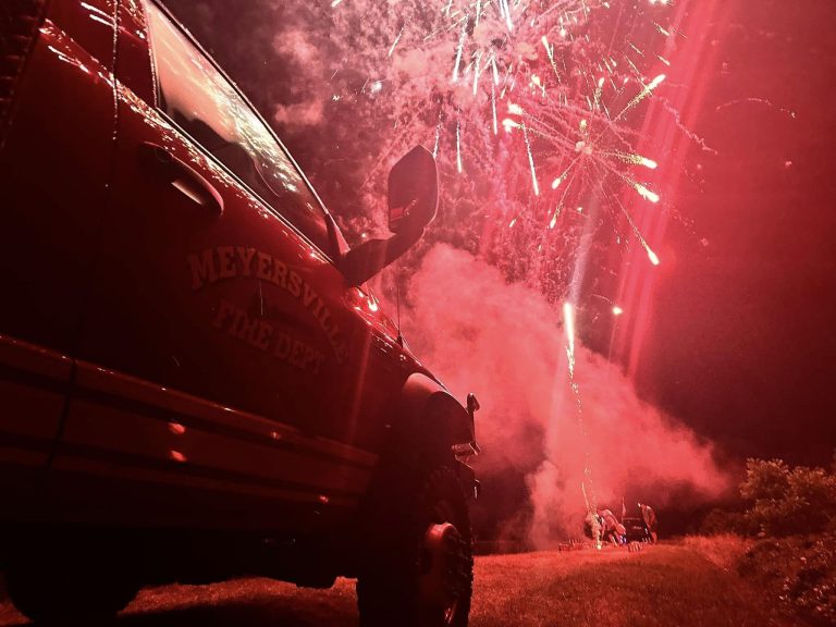 A fire department vehicle with "Meyersville Fire Dept" on the side faces a field where fireworks light up the night sky, with red hues illuminating the scene. Smoke billows near the ground and figures can be seen in the background watching the display.
