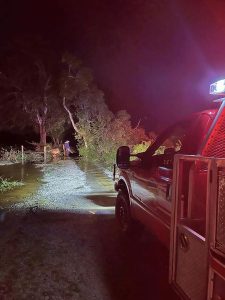 A red emergency vehicle with its door open and lights on is parked on a gravel path at night. A flooded area is ahead, with water covering part of the path. A person stands near the water, beside trees and vegetation.