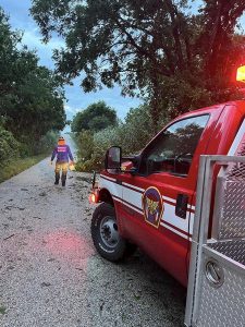 A firefighter in gear walks along a tree-lined road covered with debris. A red fire truck with its lights on is parked nearby. The sky is cloudy and the scene suggests recent storm activity.