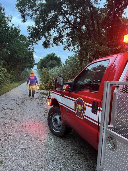 A firefighter in gear walks along a tree-lined road covered with debris. A red fire truck with its lights on is parked nearby. The sky is cloudy and the scene suggests recent storm activity.