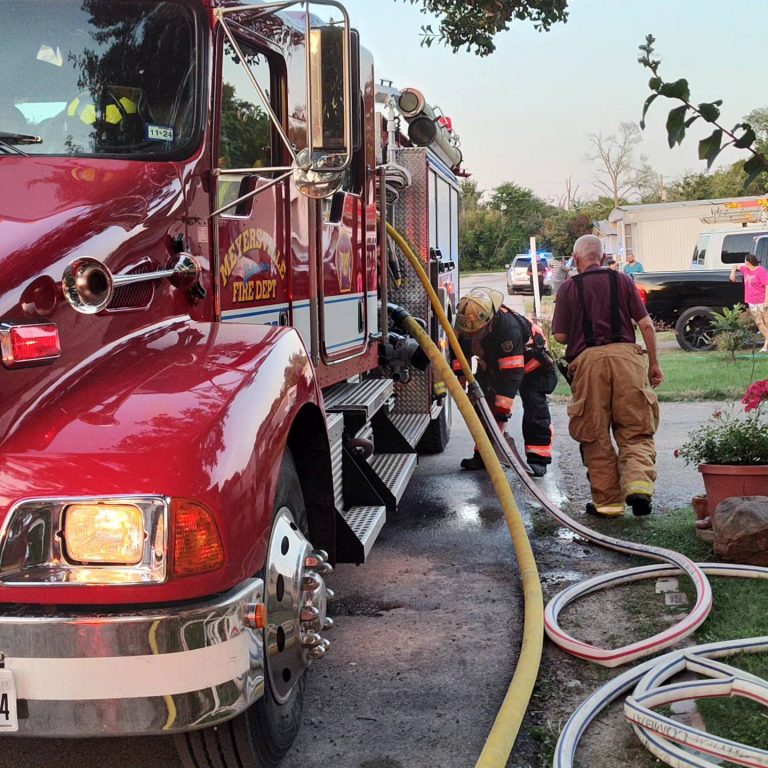 Firefighters work with hoses connected to a red firetruck labeled "Western Fire Dept" parked on a street. One firefighter stands near the truck, while another crouches to manage the hoses. Nearby, plants and garden decorations are visible in a residential area.