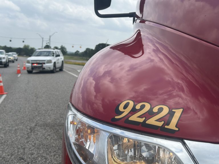 The image shows a red vehicle with the number 9221, focusing on the front headlight and part of the hood. In the background, multiple vehicles, including a white truck, are on a road with orange traffic cones and traffic lights visible under a cloudy sky.