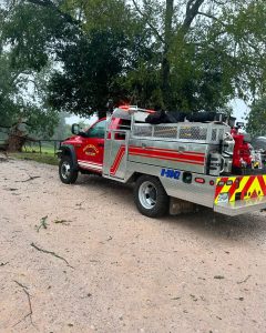 A red fire truck from the Winchester Fire Department is parked on a gravel road near a tree. The truck has emergency lights on and is equipped with various gear. Branches and debris are scattered on the ground around the vehicle.