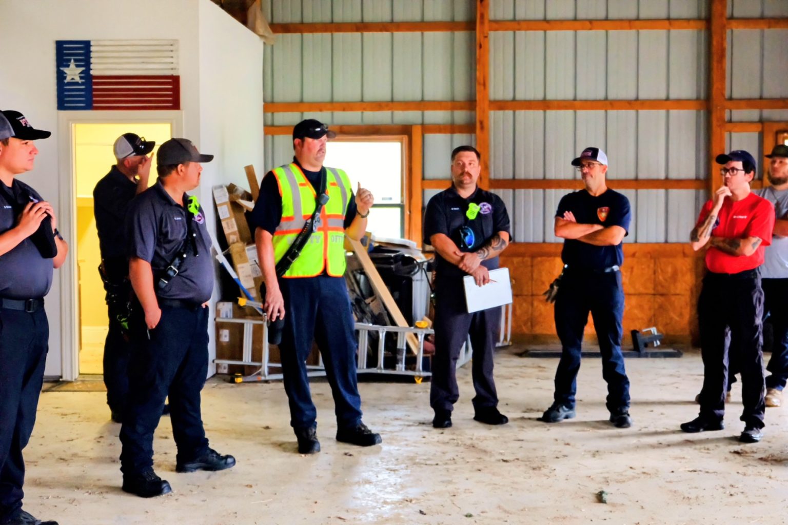 A group of firefighters in uniform stand in a semi-circle inside a garage or warehouse. One firefighter in a fluorescent safety vest appears to be addressing the others. The room has concrete flooring, various tools, and an American flag decoration on the wall.