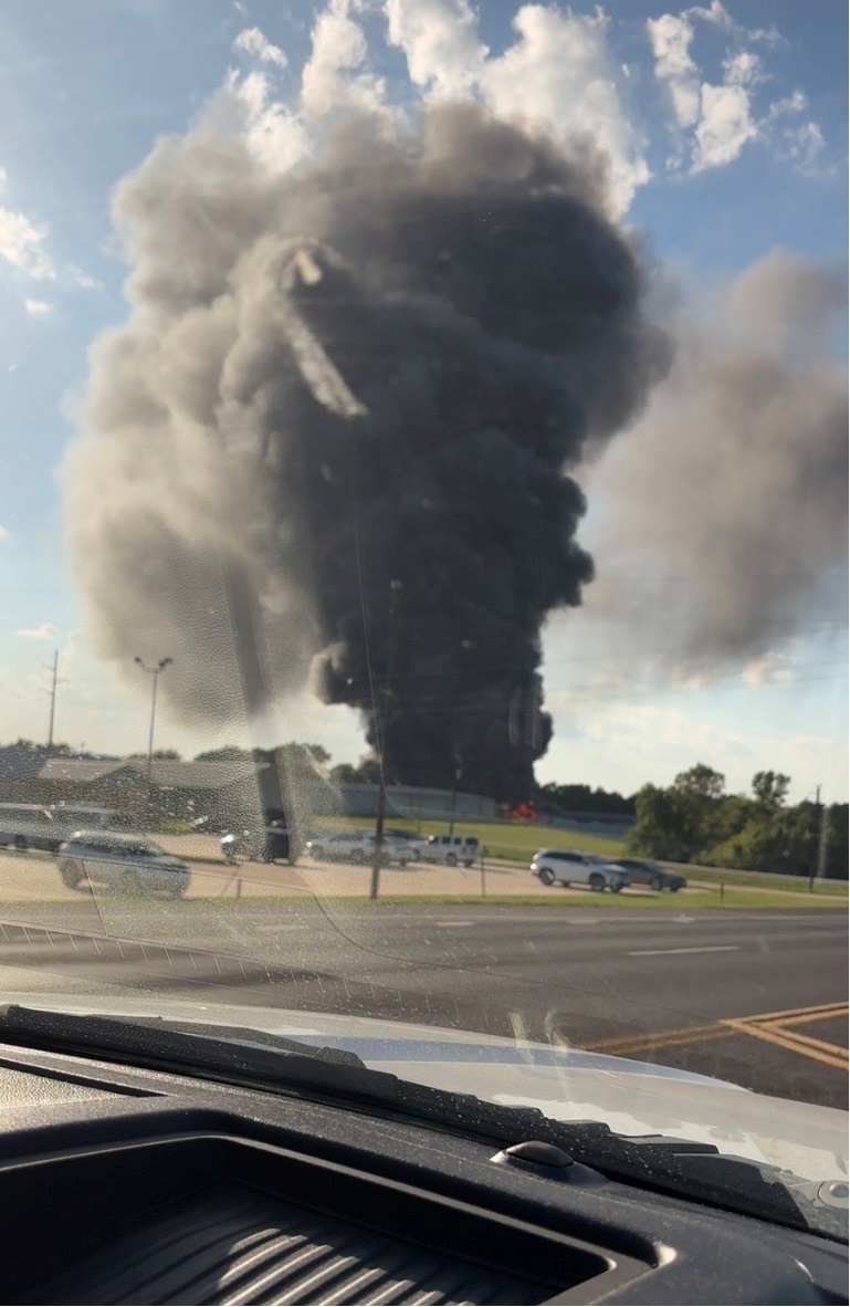A massive plume of dark smoke rises into the sky from a fire in the distance, as viewed from inside a vehicle. Cars are seen driving on the road in the foreground, and a partly cloudy sky serves as the backdrop.