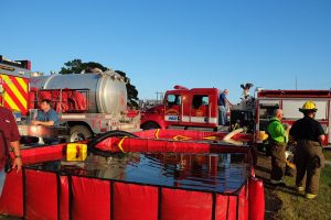 Firefighters setting up a portable water tank next to fire trucks and equipment. One person in a blue shirt operates a hose, while others in protective gear observe. The scene is set outdoors with a clear blue sky and trees in the background.