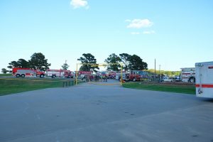A group of firefighters gathers near multiple fire trucks in an open area with trees and a blue sky in the background. They appear to be organizing equipment or preparing for an operation. The scene is calm, with no visible signs of an active emergency.