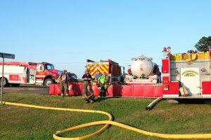Firefighters are gathered around portable water tanks and fire trucks on a grassy area beside a road. They seem to be preparing equipment, with several hoses laid out on the ground. The scene is well-lit with a clear sky overhead.