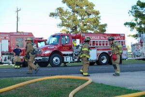 Four firefighters in uniform are in front of two red fire trucks from the Bellville Volunteer Fire Department. The scene appears to be outdoors, on a sunny day, with trees in the background. Fire hoses are laid out on the ground.
