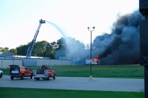 Two firefighting vehicles are parked on a paved area close to a grass field and a structure on fire. Thick black smoke rises as a large firefighter ladder truck sprays water onto the burning building. Trees are visible in the background under a clear sky.