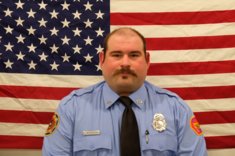 A man in a blue uniform with patches on the sleeves and a silver badge on the chest stands in front of a large American flag. He has a mustache and is wearing a black tie.