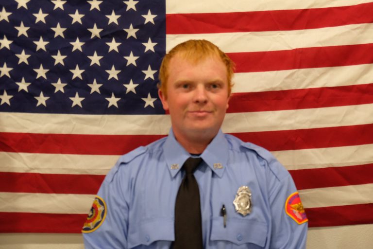 A person with red hair is wearing a blue uniform with badges and a black tie, standing in front of an American flag.