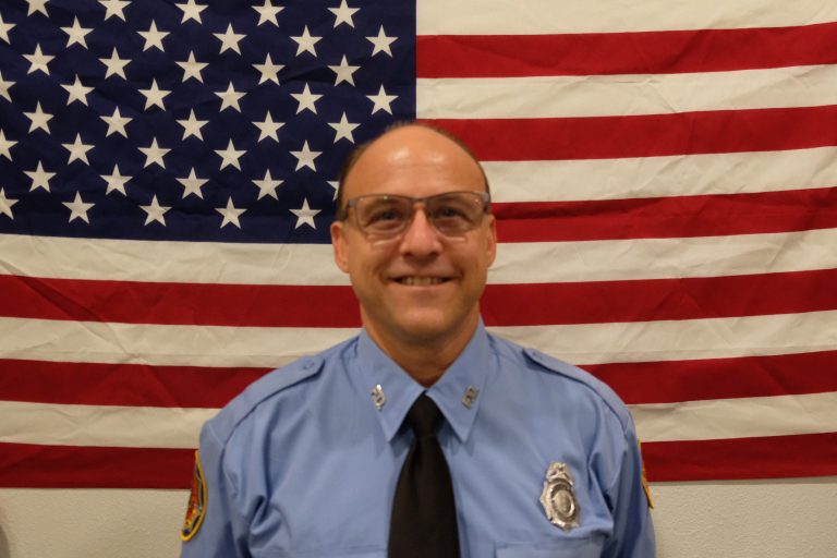A man in a blue uniform with a badge and tie stands smiling in front of an American flag. He is wearing glasses and has a badge on his shirt.