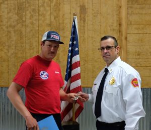 Two men stand indoors. One wears a red T-shirt and cap, holding a certificate and small silver item. The other is in a white uniform with a red patch, holding part of the item. They stand in front of a U.S. flag and a wood-paneled wall.
