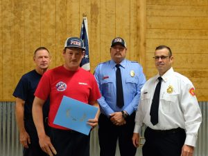 Four men, three in uniform and one holding a blue folder, stand side by side indoors. An American flag is in the background. The man with the folder wears a fire department hat. Two others wear fire department uniforms. Wood-paneled walls surround them.