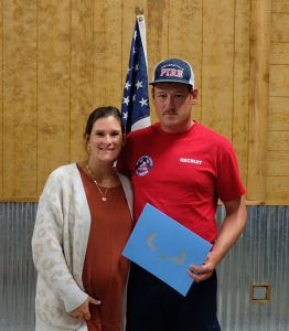 A man in a red shirt and hat, holding a blue folder, stands next to a woman wearing a beige cardigan. An American flag is in the background. Both individuals are smiling, and the setting appears to be indoors with a wooden and metal wall.