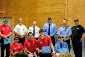 A group of firefighters poses indoors for a photo. Four are kneeling in front holding certificates. Others stand behind them in uniforms, smiling. The background features wooden walls and a door.