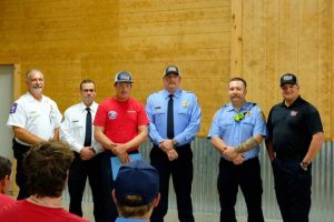 A group of six firefighters stand indoors, dressed in uniforms. Some wear blue shirts and caps, while others are in white shirts. They are posing for a photo next to a beige wall, in front of seated attendees.