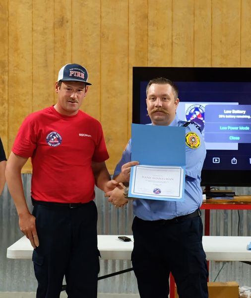 Two men stand indoors holding a certificate. One is wearing a red shirt and cap, the other is in a blue uniform. They appear to be at some sort of presentation or ceremony. A screen is visible in the background.