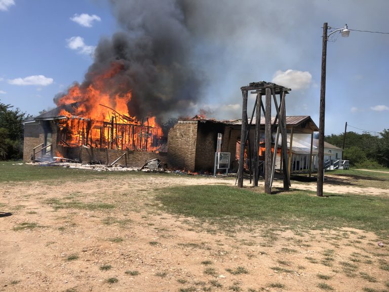 A wooden building engulfed in flames, with thick black smoke rising into a clear blue sky. The structure's walls are visible, partially collapsed, and surrounded by grass. An electricity pole stands nearby.