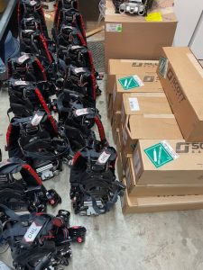 A group of black and red safety harnesses are arranged on the floor next to several stacked cardboard boxes labeled "Scott Safety." The scene appears to be in a storage or equipment room.