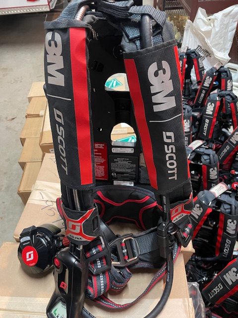 The image shows a 3M Scott harness with protective padding and adjustable straps. It includes a device with a red knob, suitable for safety use. Multiple similar harnesses are stacked in the background.