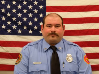 A man in a blue uniform with a badge stands in front of a large American flag. He has a mustache and is wearing a black tie.